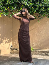 Woman in a long brown dress standing outdoors with greenery in the background
