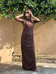 Woman in a long brown dress standing outdoors with greenery in the background
